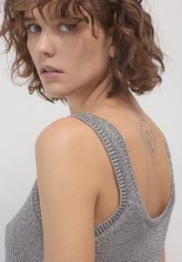 Femme aux cheveux bruns bouclés portant un haut en maille argenté sans manches, montrant un léger contour d'un tatouage sur le haut de son dos, regardant par-dessus son épaule.