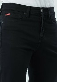 Jean en denim noir avec un design classique à cinq poches, fermeture éclair, bouton en métal, et une petite étiquette logo rouge sur la poche avant.