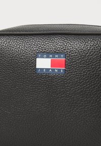 Zwarte leren tas met textuur, voorzien van een rechthoekig logo-embleem met blauwe, witte en rode elementen, geplaatst nabij de bovenrand.