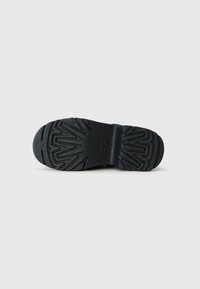 Zwarte zool van een UGG-boot met diepe zigzagprofielen en het logo zichtbaar in het midden, gefotografeerd tegen een eenvoudige witte achtergrond.