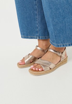 Pieds chaussés de sandales métalliques à brides croisées avec boucle à la cheville et semelle tissée style espadrilles, associées à un jean bleu raccourci.