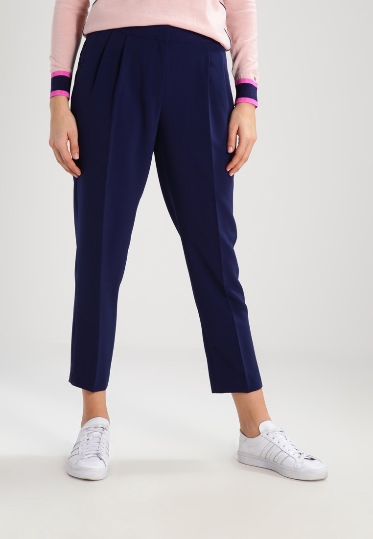 Personne portant un pantalon bleu marine ajusté longueur cheville, des baskets blanches et un pull rose clair avec des poignets bleu marine et rose, debout sur un fond uni.