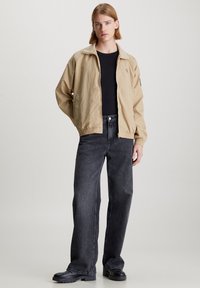 Chaqueta ligera beige con cuello alzado, puños elásticos y bolsillos laterales, llevada sobre una camisa negra y combinada con unos vaqueros de pierna ancha gris oscuro.