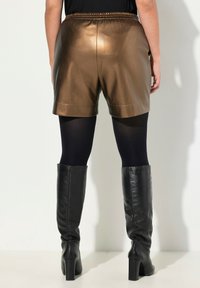Korte broeken van bruin nepleer met een elastische tailleband en een gladde textuur, gecombineerd met zwarte ondoorzichtige panty's en zwarte kniehoge laarzen.