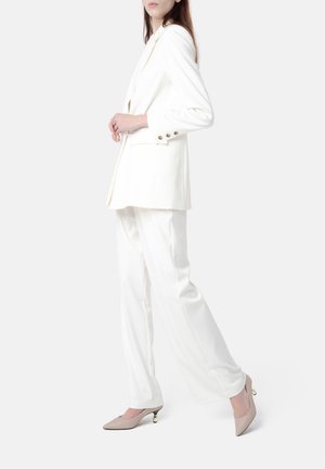 Witte blazer met inkepingen en gouden knopen, gecombineerd met witte, wijde broeken. Beige puntige hakken met gouden accenten.