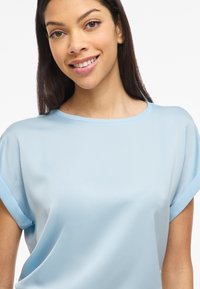 Ung kvinde med langt mørkt hår, smilende, iført en lyseblå kortærmet top foran en enkel hvid baggrund.