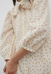 Blommig skjorta i ljus krämfärg med persikofärgade blommor, volangkrage, puffärmar och knäppning fram; tillverkad av lättviktsmaterial.