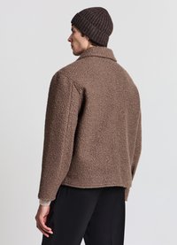 Giacca marrone testurizzata realizzata in materiale peloso, con colletto e vestibilità comoda, abbinata a un berretto marrone a coste e pantaloni neri.