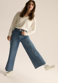 Jasnokremowy top z długim rękawem połączony z niebieskimi spodniami dżinsowymi o średnim stanie i białymi trampkami. Dżinsy posiadają klasyczne zapięcie na guzik i zamek.