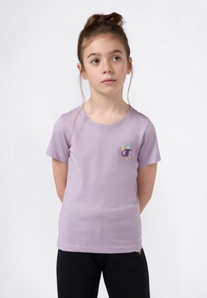 Jeune fille aux cheveux bruns noués, portant un t-shirt violet clair avec un petit graphique coloré et un pantalon noir, se tenant les mains derrière le dos.