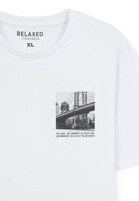 T-shirt bianca in cotone con una grafica quadrata in bianco e nero di un ponte e dello skyline di una città. Il testo sotto l'immagine descrive luci e grattacieli.