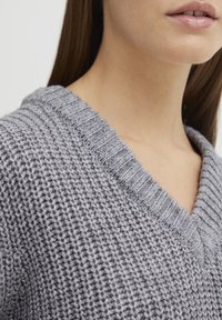 Grauer Strickpullover mit V-Ausschnitt, aus strukturiertem Stoff und mit geripptem Kragen. Die Nahaufnahme hebt die Webdetails hervor.