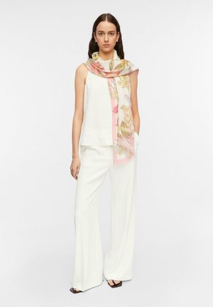 Woman wearing a sleeveless white top and wide-leg white pants, with a pink and beige floral scarf draped around her neck.