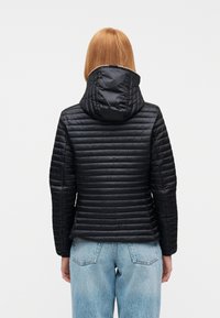 Γυναίκα με ίσια κόκκινα μαλλιά, που φοράει μαύρο καπιτονέ μπουφάν με κουκούλα και ανοιχτό μπλε τζιν, εμφανίζεται από πίσω σε λευκό φόντο.