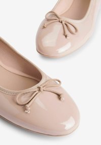 Vaaleanpunaiset patenttiballet-tyyliset tasapohjakengät, joissa on pyöreä kärki, elastinen reunus ja pieni nauha edessä. Sujuva pinta, minimalistinen muotoilu.