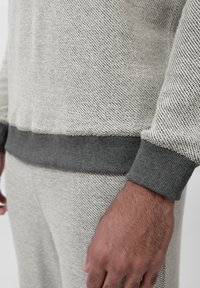 Lys grå strikket sweater med et struktureret mønster, der har mørkegrå ribbede manchetter. Nærbillede af ærmedetalje og arm.