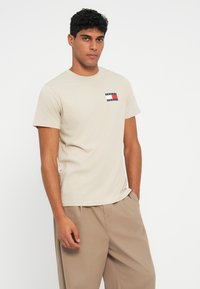 Μπεζ βαμβακερή μπλούζα με κοντό μανίκι και μικρό λογότυπο σε κόκκινο, λευκό και μπλε. Απλή στρογγυλή λαιμόκοψη, κοντά μανίκια και χαλαρή εφαρμογή.