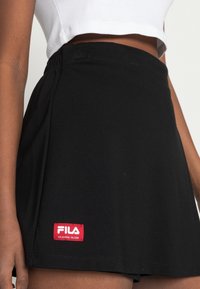 Černá sportovní sukně s pružným pasem a výraznou červenou nášivkou Fila. Hladká textura s rozšířeným designem.