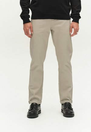 Hombre con pantalones beige de pierna recta con dobladillos enrollados, suéter negro y zapatos de cuero negros de pie sobre un fondo blanco.