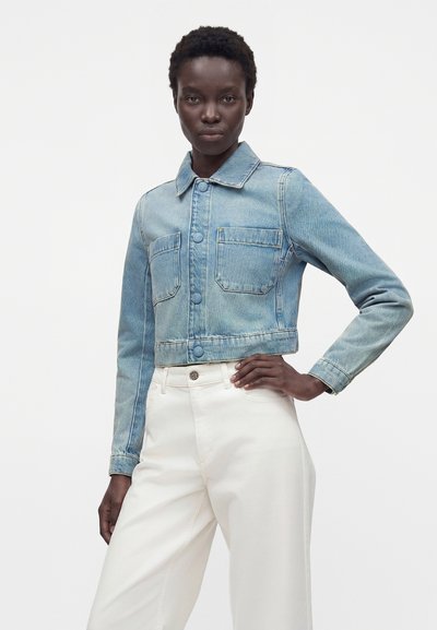 Μαύρη γυναίκα που φορά ένα ανοιχτού μπλε cropped denim μπουφάν με εμπρόσθιες τσέπες και λευκό ψηλόμεσο παντελόνι, στέκεται με το ένα χέρι στη μέση.