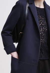 Navy wool blend coat with a structured design, notched collar, visible stitching, and large pockets. Layered with black lace top underneath.
