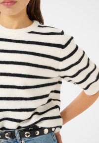 Pull en mélange de laine blanche avec des rayures horizontales noires. Il présente un col rond et des manches courtes, associé à une ceinture noire cloutée.