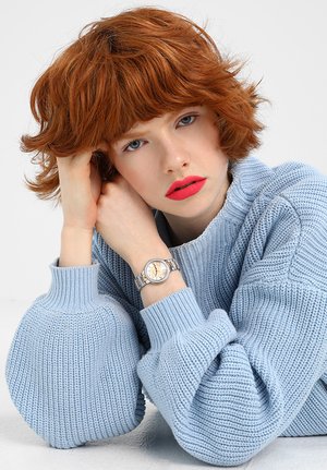 Persona joven con cabello corto y rojo y labios pintados de un rojo brillante, que lleva un suéter de punto azul claro y un reloj de pulsera plateado, apoyando la cabeza en la mano.