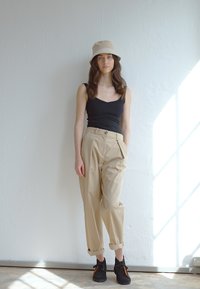Schwarzes Tanktop, beige Hochbundhose mit Falten und schwarze Schuhe mit orangefarbenen Akzenten. Beiger Bucket Hat, strukturierter Hintergrund.
