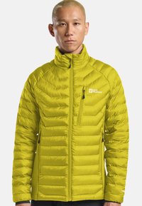 Persona che indossa un giubbotto trapuntato Jack Wolfskin verde lime con tasche con zip e colletto alto, in piedi di fronte a uno sfondo chiaro e uniforme.