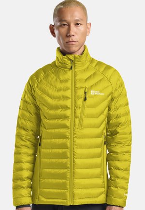 Persoon in een limoen groene gewatteerde Jack Wolfskin jas met ritssluitingen en hoge kraag, staand tegen een effen lichte achtergrond.