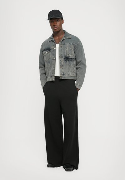 Jachetă din denim gri cu o croială lejeră, având două buzunare pe piept și închideri cu nasturi, asortată cu pantaloni negri cu croială largă și o șapcă neagră.