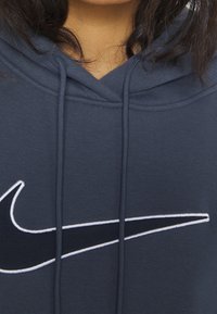 Un sweat à capuche bleu marine en mélange de coton, arborant un grand logo Nike swoosh noir entouré de blanc, avec des cordons de serrage au niveau du col.