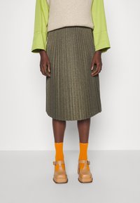 Gonna plissettata verde oliva con finitura metallica, abbinata a calzini arancioni alla caviglia e scarpe chunky beige con cinturini. Maglione in misto lana di un beige chiaro.