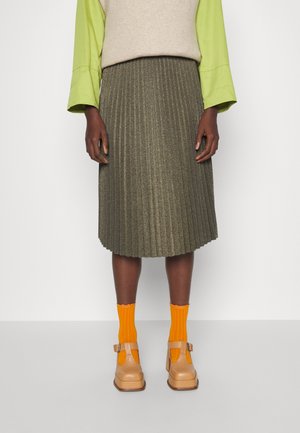 Jupe plissée vert olive avec un éclat métallique, associée à des chaussettes de cheville orange et des chaussures beiges à bouts larges et à brides. Pull en mélange de laine de couleur beige clair.