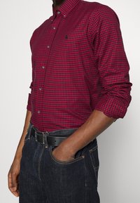 Rood en zwart geruit overhemd met lange mouwen, kraag en zilveren knopen, gecombineerd met donkere spijkerbroek en een zwarte riem.