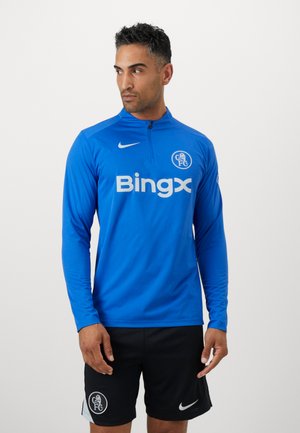 Blå langærmet sportstrøje med en kvart-zip, der har et hvidt logo og tekst på fronten. Båret med sorte shorts og en rund logodetalje.