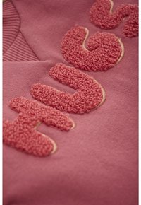 Rosa Stoff mit erhöhten, strukturierten Buchstaben aus einem flauschigen, chenille-ähnlichen Material, das auf die Oberfläche gestickt ist.