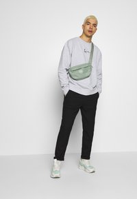 Grøn crossbody taske med lynlåslukning, båret over en lys grå sweatshirt og sorte bukser, parret med hvide sneakers med mint detaljer.
