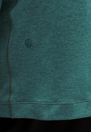 Material verde teal de echipament sportiv cu cusături vizibile și un mic logo circular pe partea stângă, aproape de tiv.