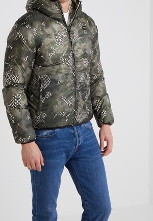 Homme portant une doudoune à capuche aux motifs camouflage verts et marron, avec un jean bleu, debout devant un fond clair uni.