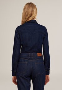 Chaqueta de mezclilla azul oscuro con un cuello clásico, detalles de botones y acentos decorativos a lo largo del cuello. Parche de marca de cuero en la parte trasera de la cinturilla.