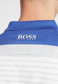 Pikétröja med en blå krage med vita ränder och en logotyp. Tyget är texturerat, med en slät ytfinish och sydda detaljer.