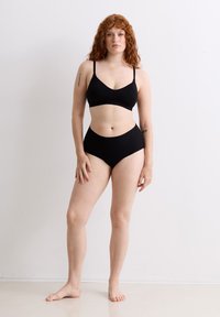 Eine rothaarige Frau mit Tattoos, die einen schwarzen BH und hochgeschnittene Unterwäsche trägt, steht barfuß vor einer schlichten weißen Wand.