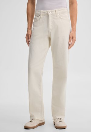 Person, die off-white Jeans mit geradem Bein, weiße Sneaker und ein weißes Hemd trägt und vor einem schlichten hellen Hintergrund steht.