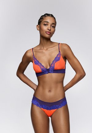Vrouw met gevlochten haar die oranje en paarse kanten lingerie draagt, staande met handen in de zij tegen een effen lichte achtergrond.