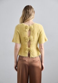Suéter de mohair amarillo con mangas cortas, que presenta tres lazos y un escote tipo ojo de cerradura en la parte posterior. Combinado con pantalones marrones con bolsillos laterales.