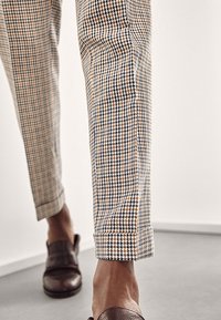 Gros plan sur des jambes portant un pantalon à motif pied-de-poule marron et blanc avec des chaussures en cuir marron sans lacets sur un sol gris.