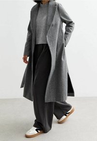 Abrigo largo de espiga gris con solapas de muesca, llevado sobre un suéter gris y pantalones anchos oscuros, combinado con zapatillas blancas con acentos negros.