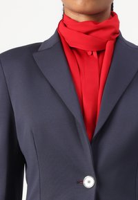 Tengerészkék blézer strukturált anyagból, egyetlen ezüst gombbal, élénk piros blúzra rétegezve, nyakkendős és gombos záródással.