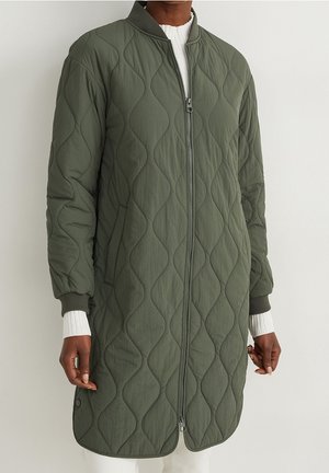 Le modèle porte un long manteau vert olive matelassé à losanges, avec une fermeture éclair sur le devant, des poignets côtelés et un pull blanc en dessous.
